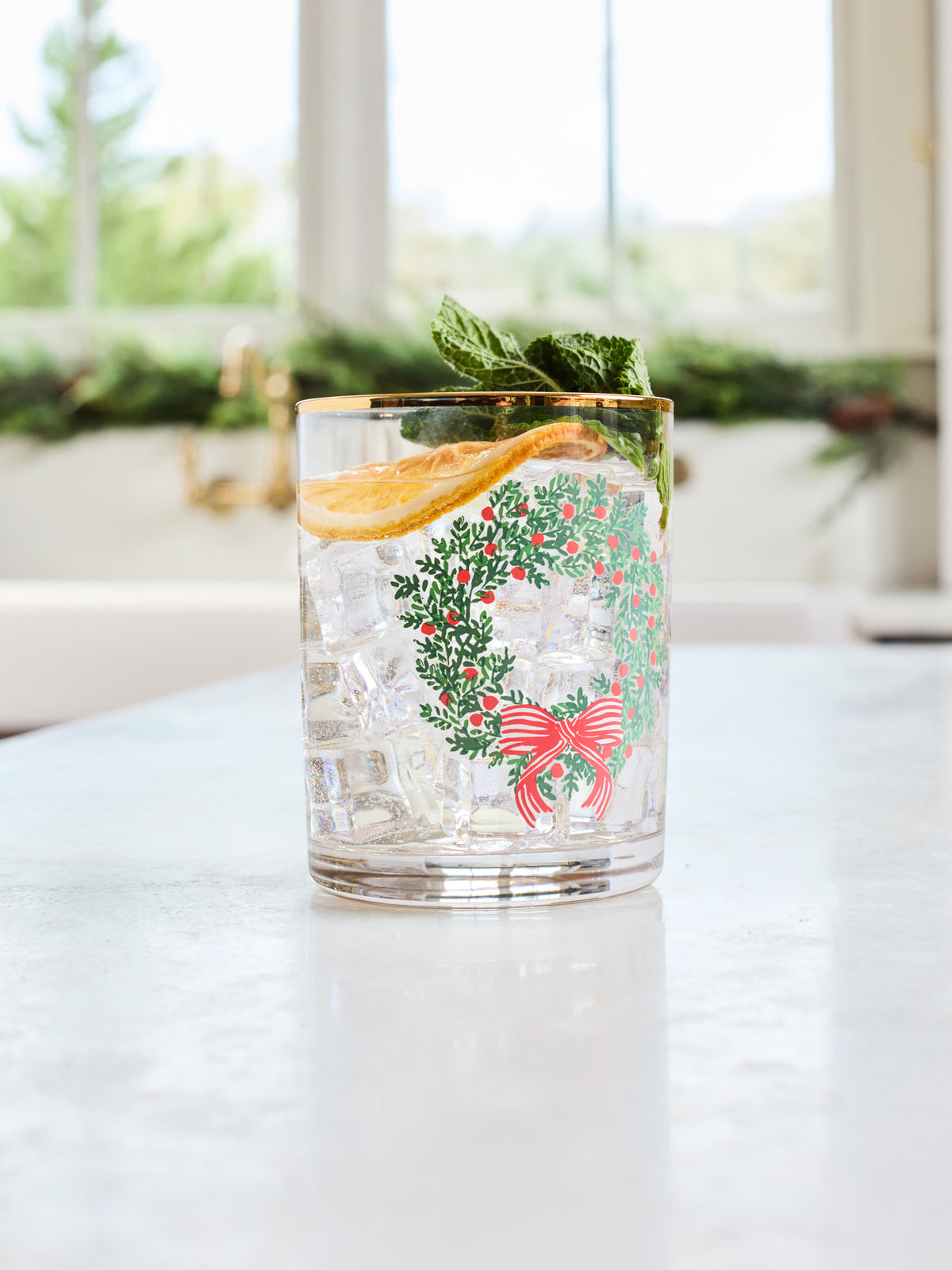 A clear glass tumbler with a gold rim and a festive green wreath and red bow print, filled with a clear cocktail and garnished with a mint leaf and orange slice, set on a light countertop with a blurred kitchen background.
