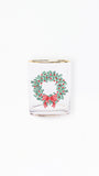 A clear glass shot glass featuring a printed green holly wreath with red berries and a red bow, finished with a gold rim, set against a plain white background.
