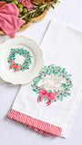 A white holiday tea towel with a green wreath and red bow print, featuring red and white striped ruffled trim, displayed next to a matching white plate on a light background.