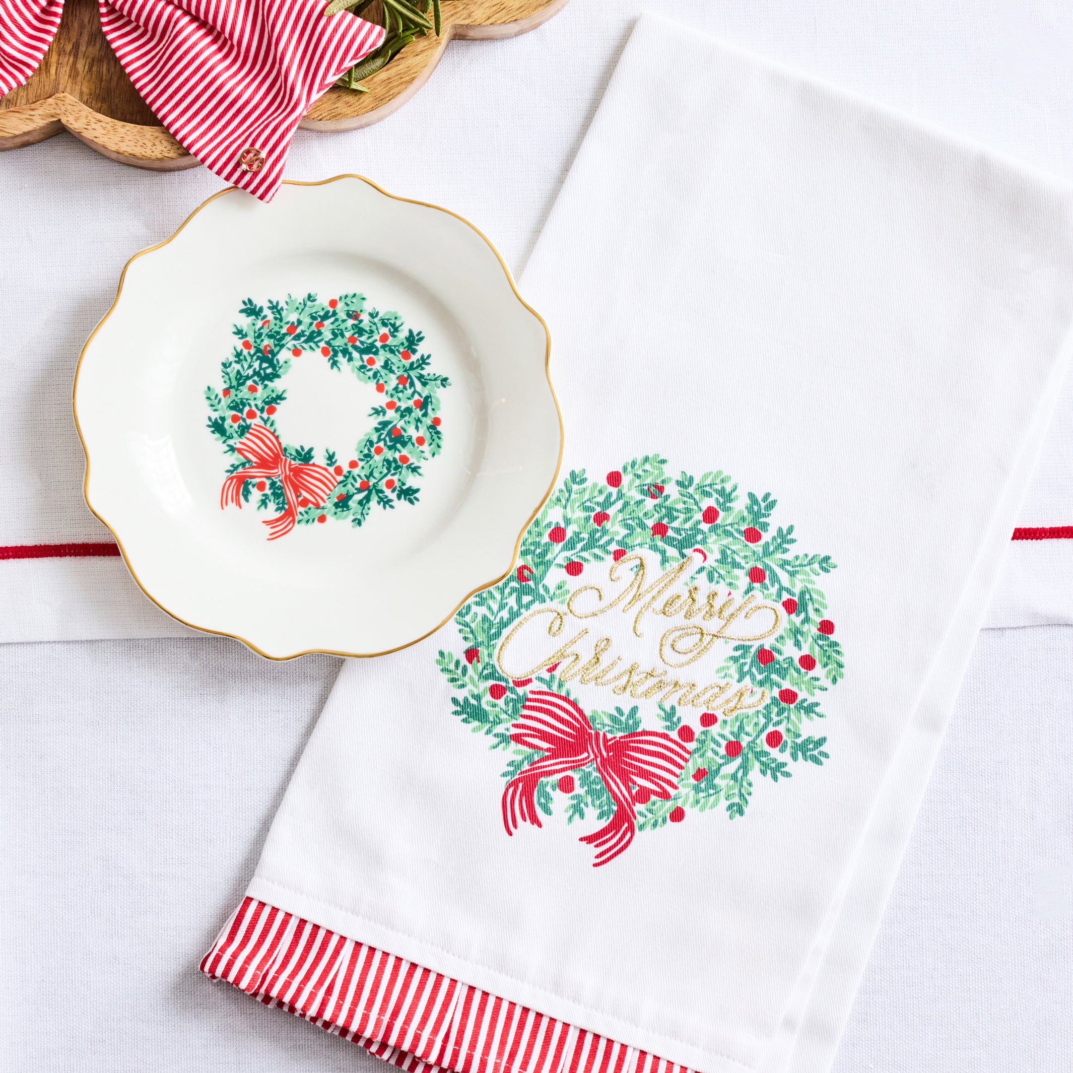 A white holiday tea towel with a green wreath and red bow print, featuring red and white striped ruffled trim, displayed next to a matching white plate on a light background.