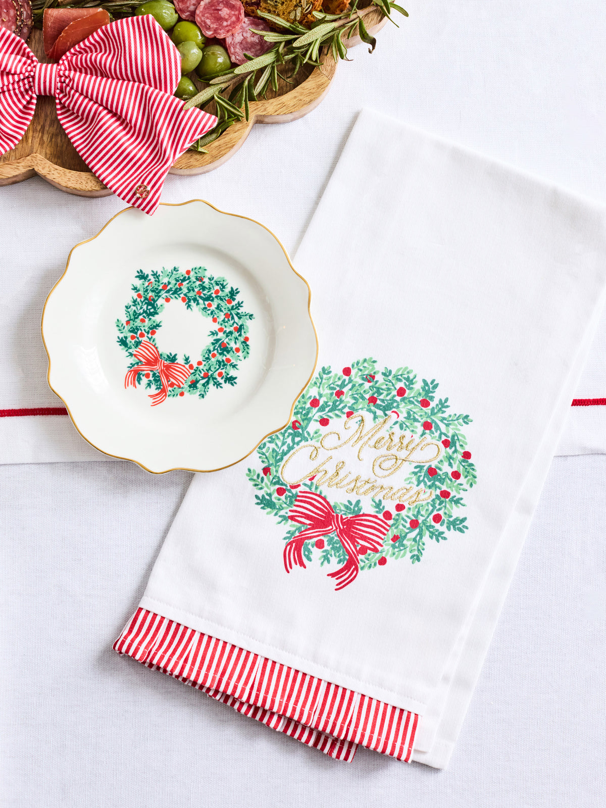 A white holiday tea towel with a green wreath and red bow print, featuring red and white striped ruffled trim, displayed next to a matching white plate on a light background.