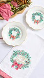 Three white ceramic holiday plates with gold trim and green wreath motifs are arranged on a white surface beside a white napkin with a large 'Merry Christmas' wreath print and a red-and-white striped napkin edge. A decorative charcuterie board is partially visible in the top left.