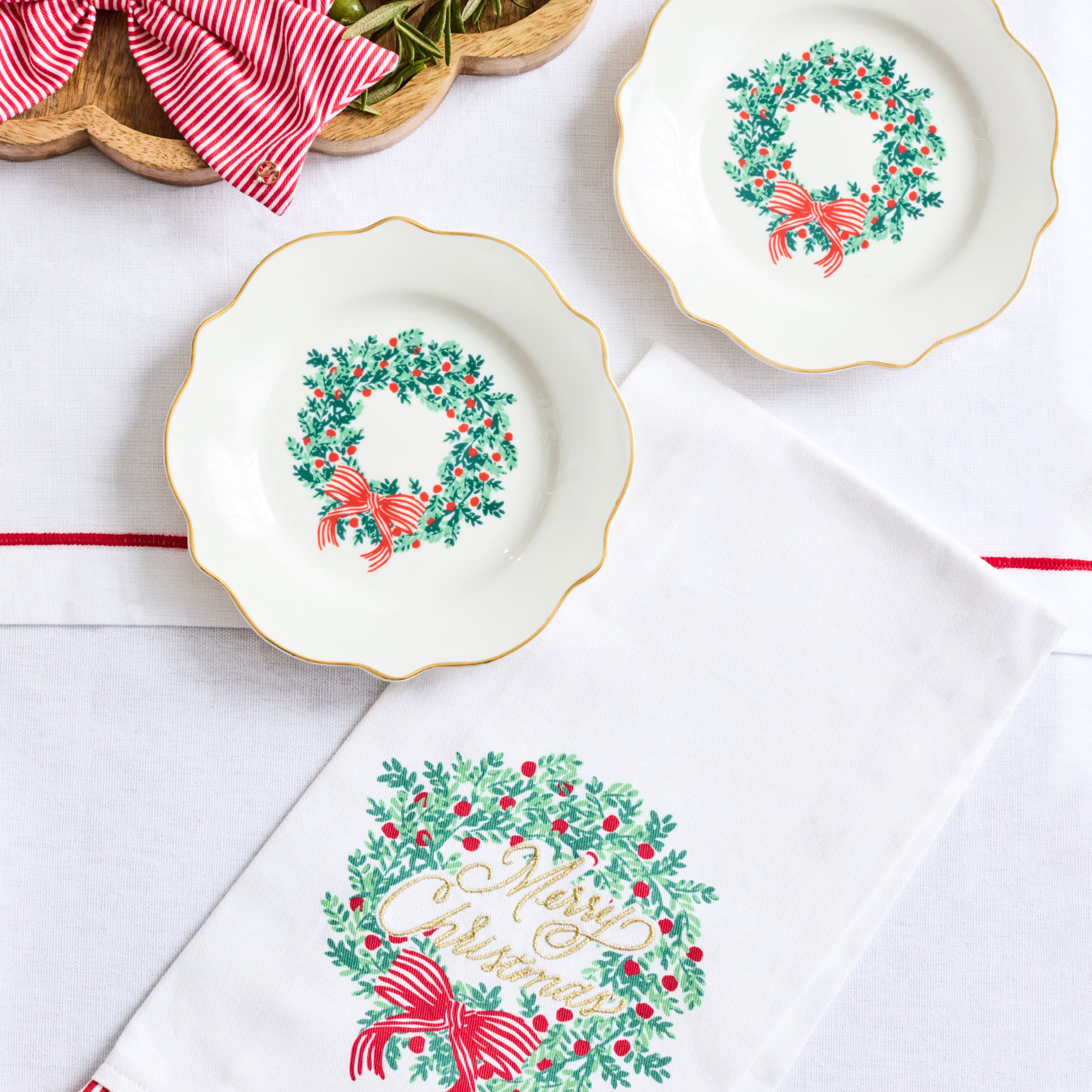 Three white ceramic holiday plates with gold trim and green wreath motifs are arranged on a white surface beside a white napkin with a large 'Merry Christmas' wreath print and a red-and-white striped napkin edge. A decorative charcuterie board is partially visible in the top left.