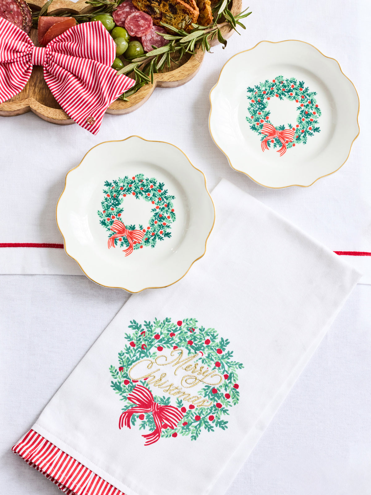 Three white ceramic holiday plates with gold trim and green wreath motifs are arranged on a white surface beside a white napkin with a large 'Merry Christmas' wreath print and a red-and-white striped napkin edge. A decorative charcuterie board is partially visible in the top left.