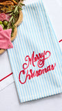 A light blue and white striped kitchen towel with 'Merry Christmas' embroidered in red script, styled on a white surface with a wooden board of appetizers and a red striped bow.
