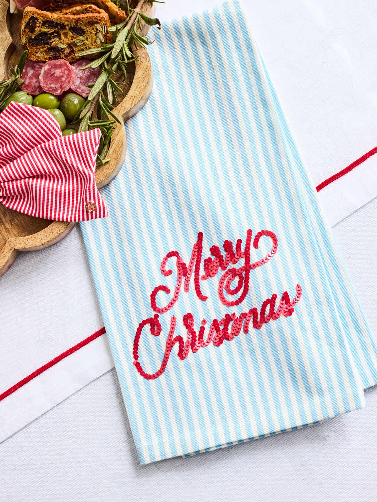A light blue and white striped kitchen towel with 'Merry Christmas' embroidered in red script, styled on a white surface with a wooden board of appetizers and a red striped bow.