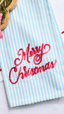 A light blue and white vertically striped kitchen towel with 'Merry Christmas' embroidered in red script across the front, styled on a white surface with holiday props.