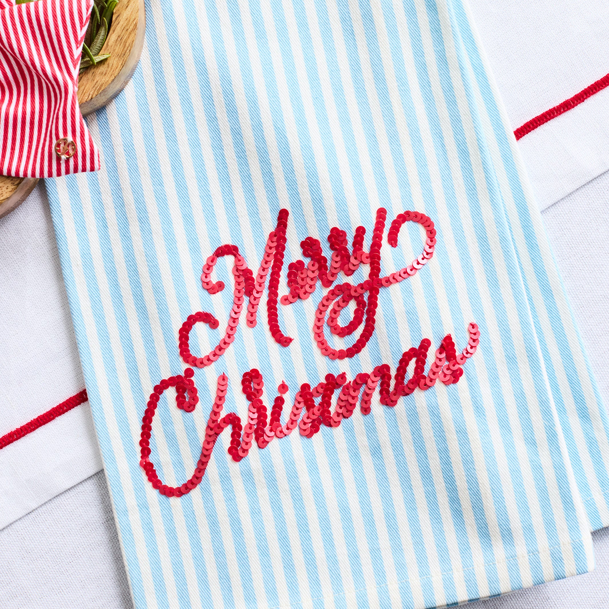 A light blue and white vertically striped kitchen towel with 'Merry Christmas' embroidered in red script across the front, styled on a white surface with holiday props.