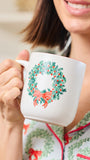 A person holding a white ceramic mug decorated with a green holly wreath and red bow, wearing matching holiday pajamas in a softly lit indoor setting.