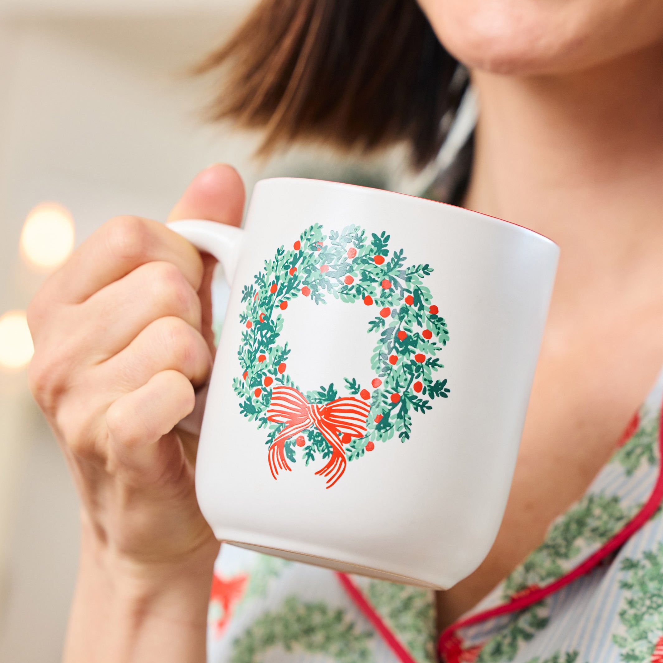 A person holding a white ceramic mug decorated with a green holly wreath and red bow, wearing matching holiday pajamas in a softly lit indoor setting.
