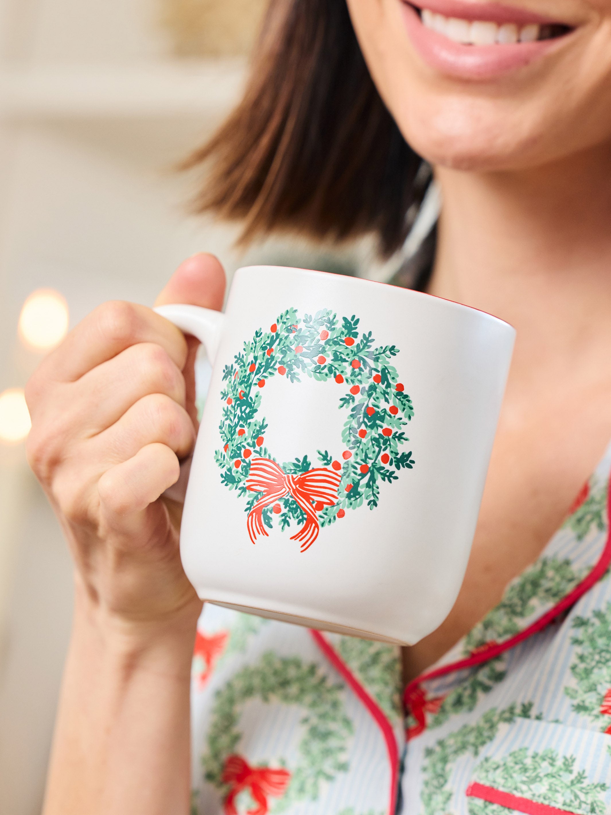 A person holding a white ceramic mug decorated with a green holly wreath and red bow, wearing matching holiday pajamas in a softly lit indoor setting.
