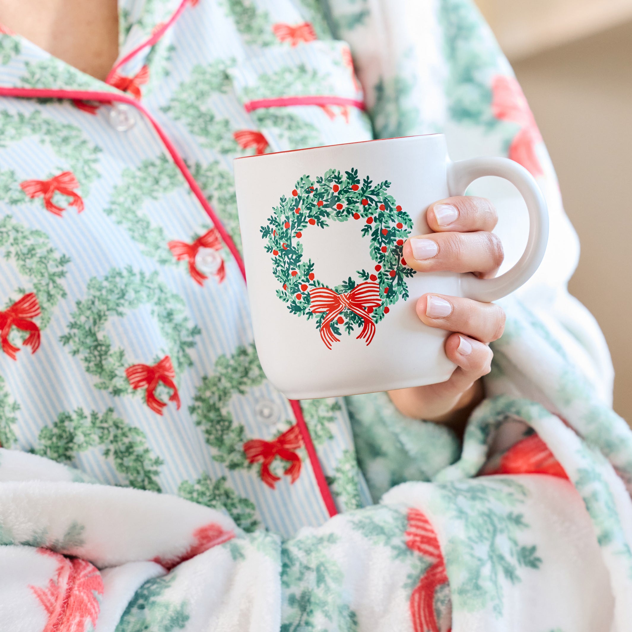 A person wearing festive holiday pajamas with a green wreath and red bow pattern, holding a white ceramic mug decorated with a matching wreath design.