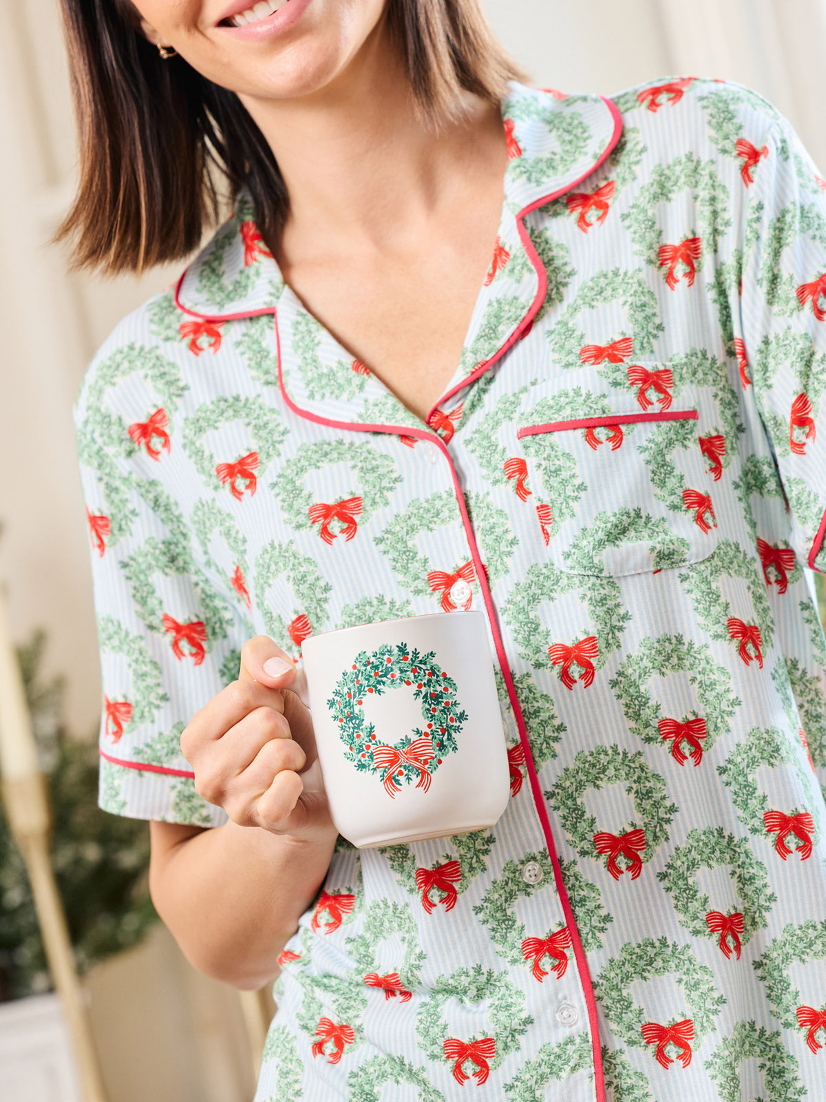 A woman wearing a short-sleeve pajama top with a green wreath and red bow holiday print, holding a white mug with a matching wreath design. The pajamas have red piping trim.