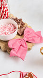 Two red and white striped fabric napkins tied into bows, displayed on a scalloped wooden board with candy-themed props including crushed candy cane pieces, chocolate bars, a jar of striped straws, and a gingerbread cookie.