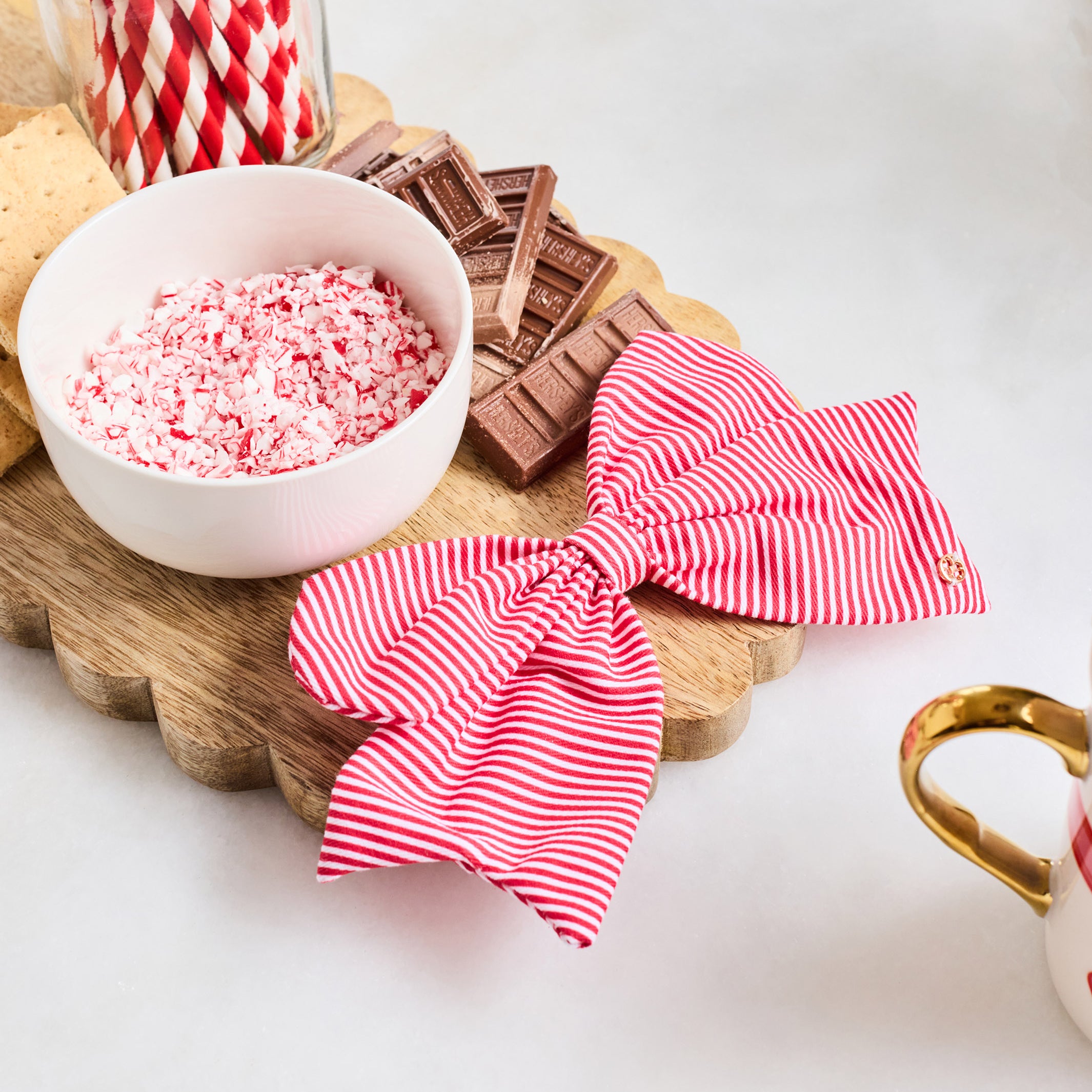Two red and white striped fabric napkins tied into bows, displayed on a scalloped wooden board with candy-themed props including crushed candy cane pieces, chocolate bars, a jar of striped straws, and a gingerbread cookie.