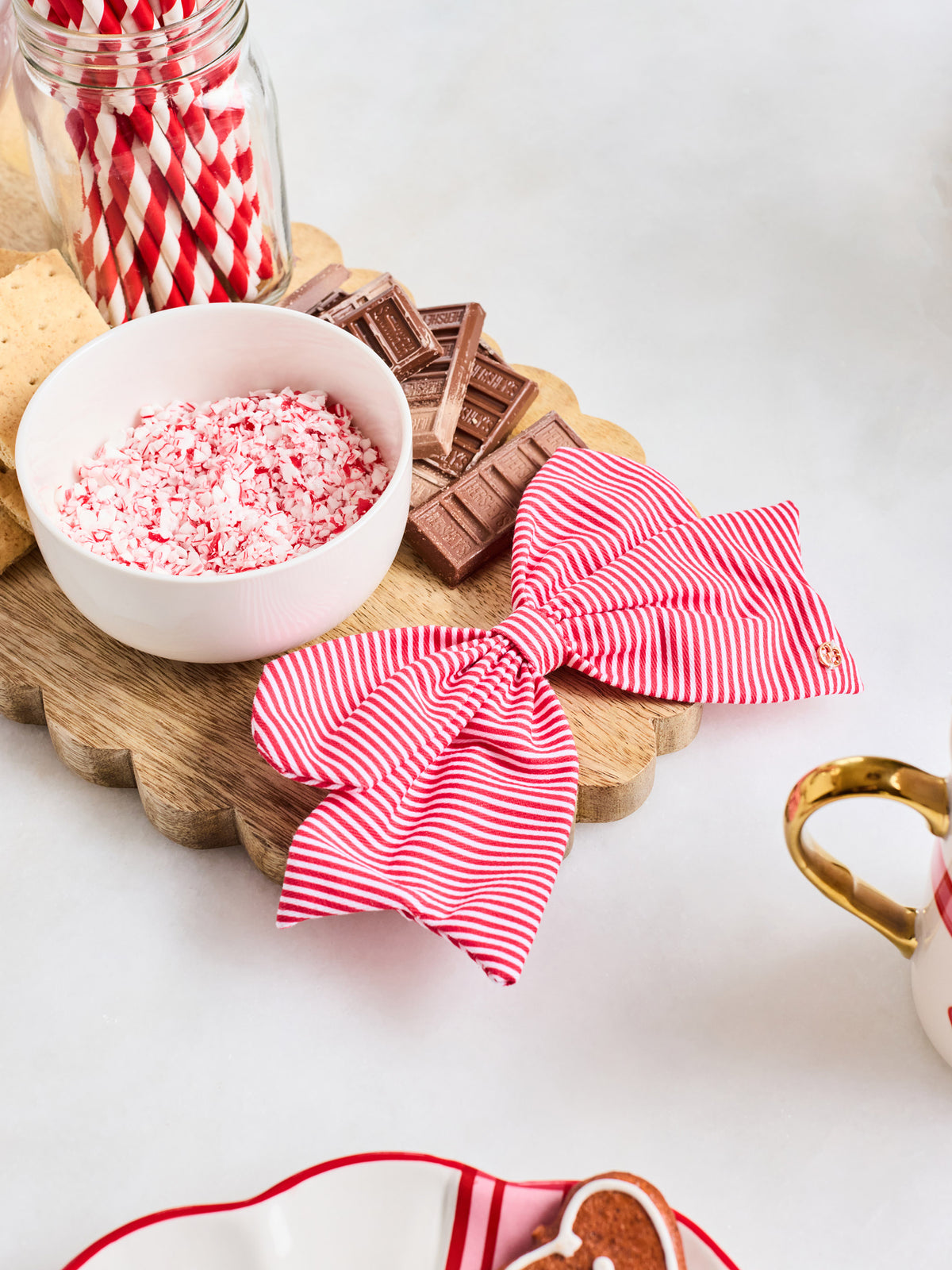Two red and white striped fabric napkins tied into bows, displayed on a scalloped wooden board with candy-themed props including crushed candy cane pieces, chocolate bars, a jar of striped straws, and a gingerbread cookie.