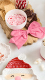 A styled holiday tabletop scene with a wooden serving board holding crackers, chocolate pieces, a bowl of crushed candy canes, a red-and-white striped fabric bow, marshmallows, and Santa-themed ceramic dishes on a white surface.
