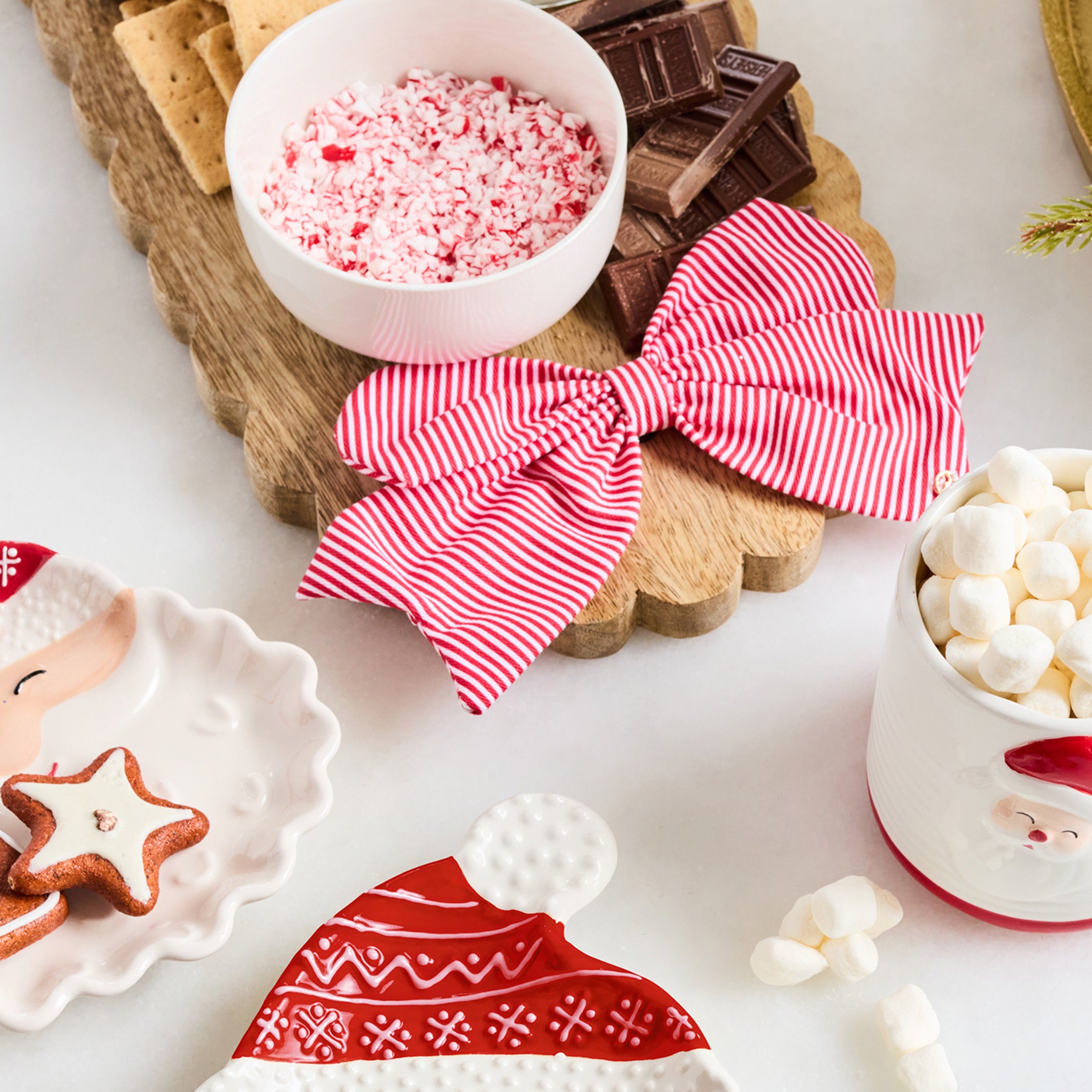 A styled holiday tabletop scene with a wooden serving board holding crackers, chocolate pieces, a bowl of crushed candy canes, a red-and-white striped fabric bow, marshmallows, and Santa-themed ceramic dishes on a white surface.