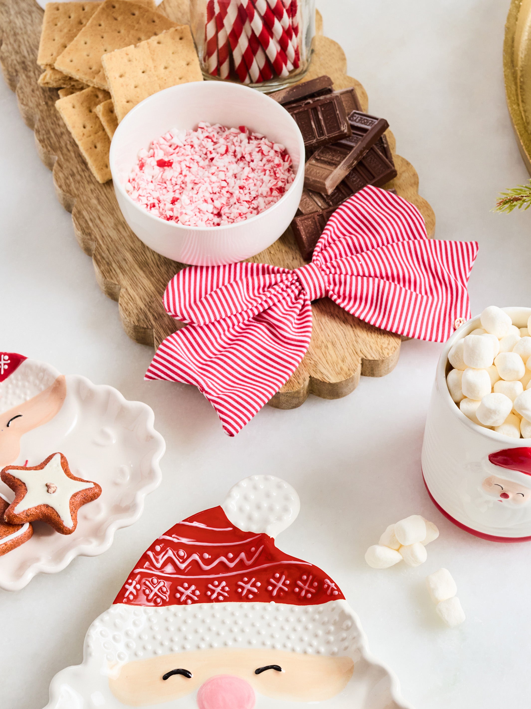 A styled holiday tabletop scene with a wooden serving board holding crackers, chocolate pieces, a bowl of crushed candy canes, a red-and-white striped fabric bow, marshmallows, and Santa-themed ceramic dishes on a white surface.