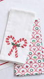 A white fabric tea towel with a red embroidered candy cane and green star motif, layered over another white towel featuring a red, green, and blue Christmas patterned border.