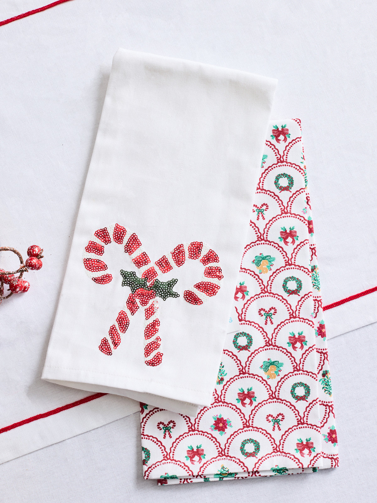 A white fabric tea towel with a red embroidered candy cane and green star motif, layered over another white towel featuring a red, green, and blue Christmas patterned border.