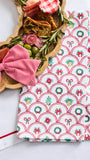 A festive kitchen towel with a red, green, and white holiday pattern is styled on a white table next to a wooden charcuterie board and a matching red striped bow.