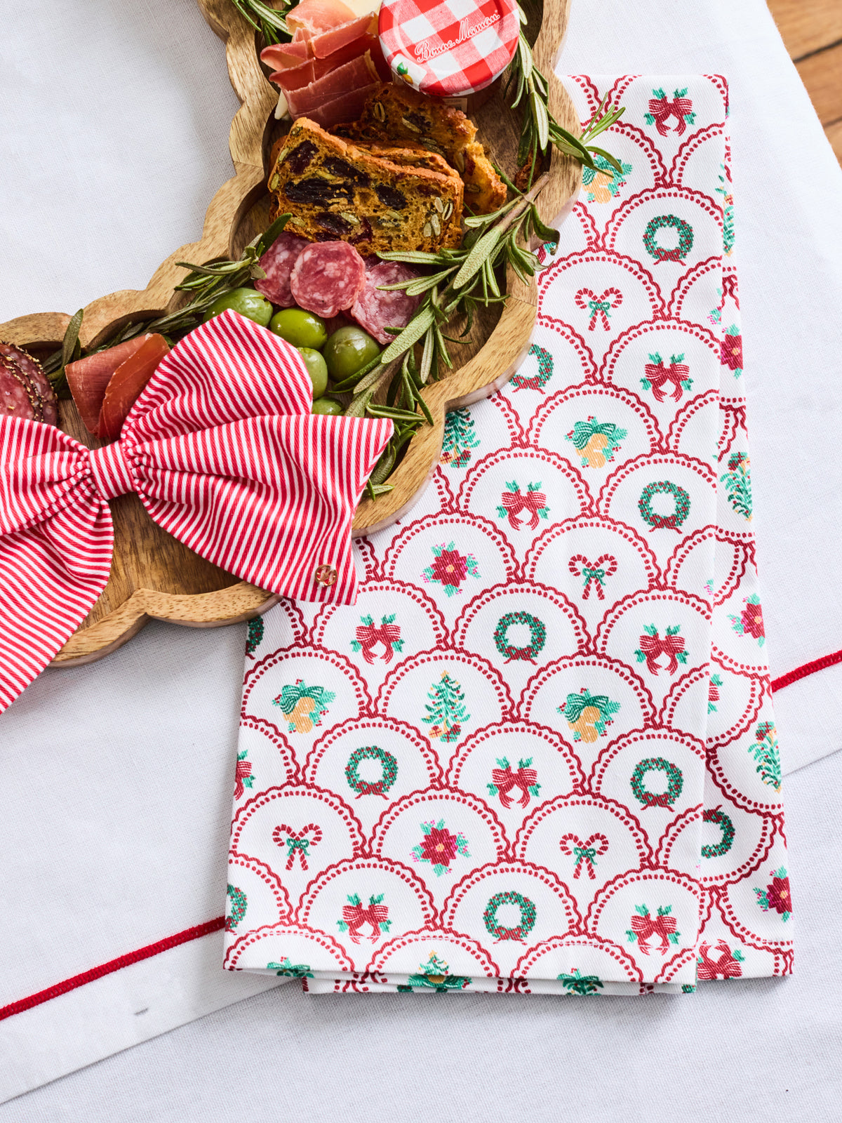 A festive kitchen towel with a red, green, and white holiday pattern is styled on a white table next to a wooden charcuterie board and a matching red striped bow.