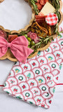 A festive holiday kitchen towel with a red and green scallop pattern and bow print, styled next to a wooden charcuterie board with cheese, salami, olives, and a red gingham checkered napkin.
