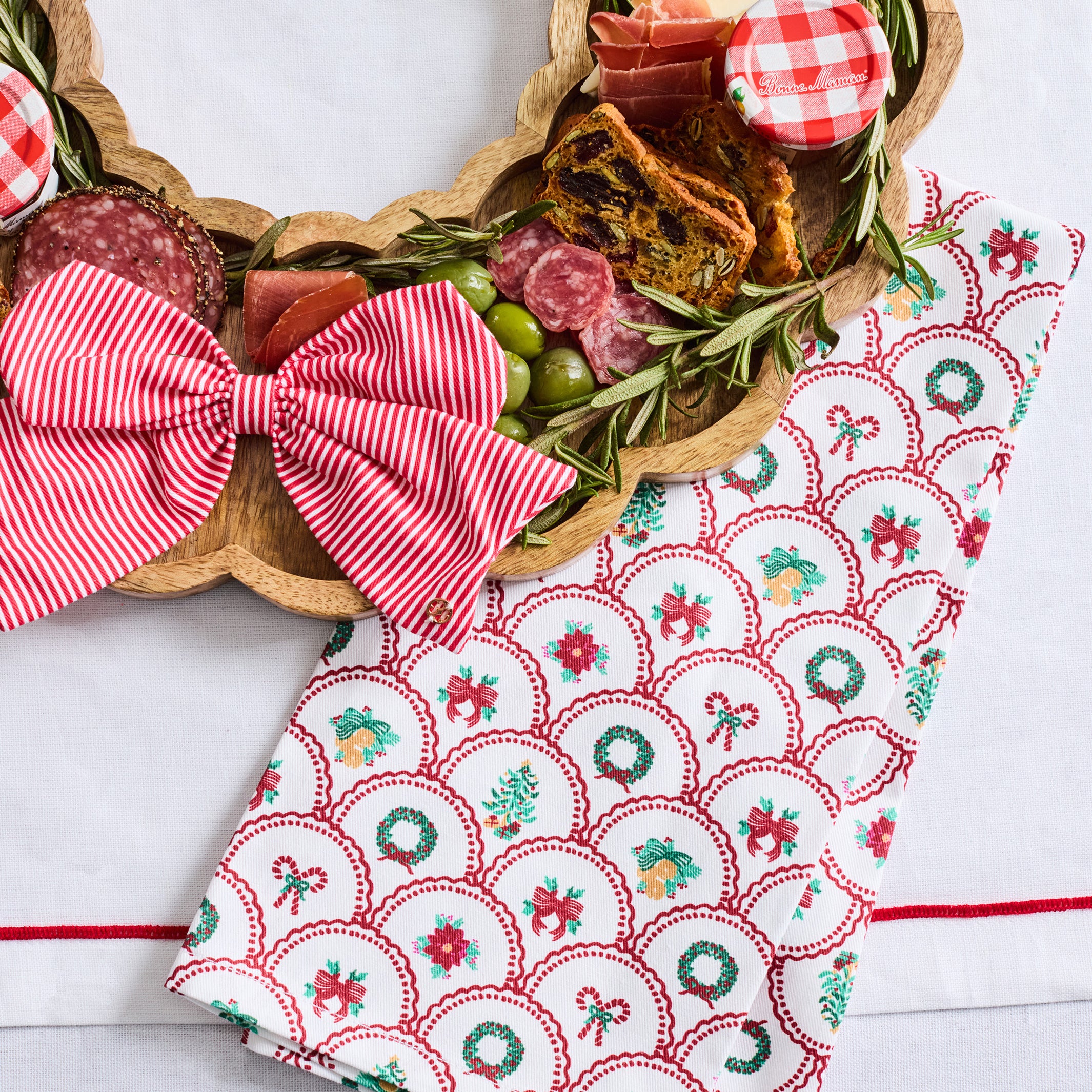 A festive holiday kitchen towel with a red and green scallop pattern and bow print, styled next to a wooden charcuterie board with cheese, salami, olives, and a red gingham checkered napkin.