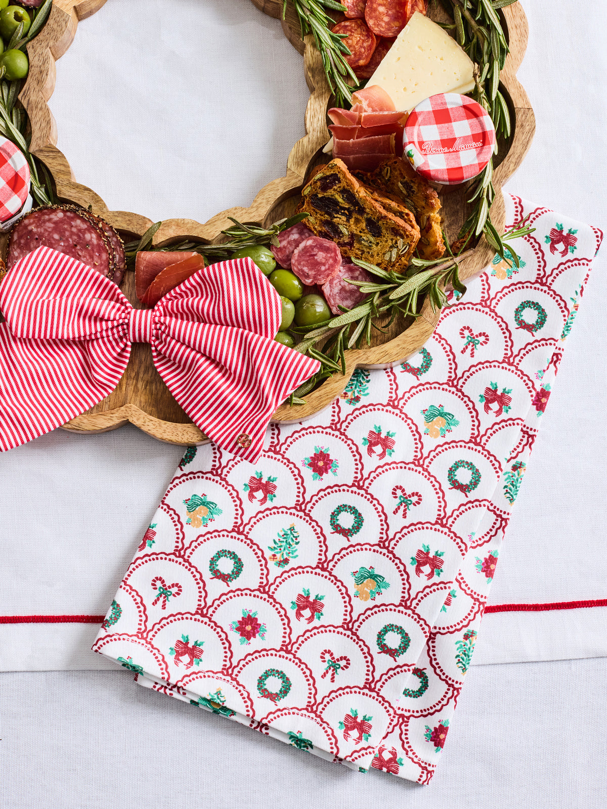 A festive holiday kitchen towel with a red and green scallop pattern and bow print, styled next to a wooden charcuterie board with cheese, salami, olives, and a red gingham checkered napkin.