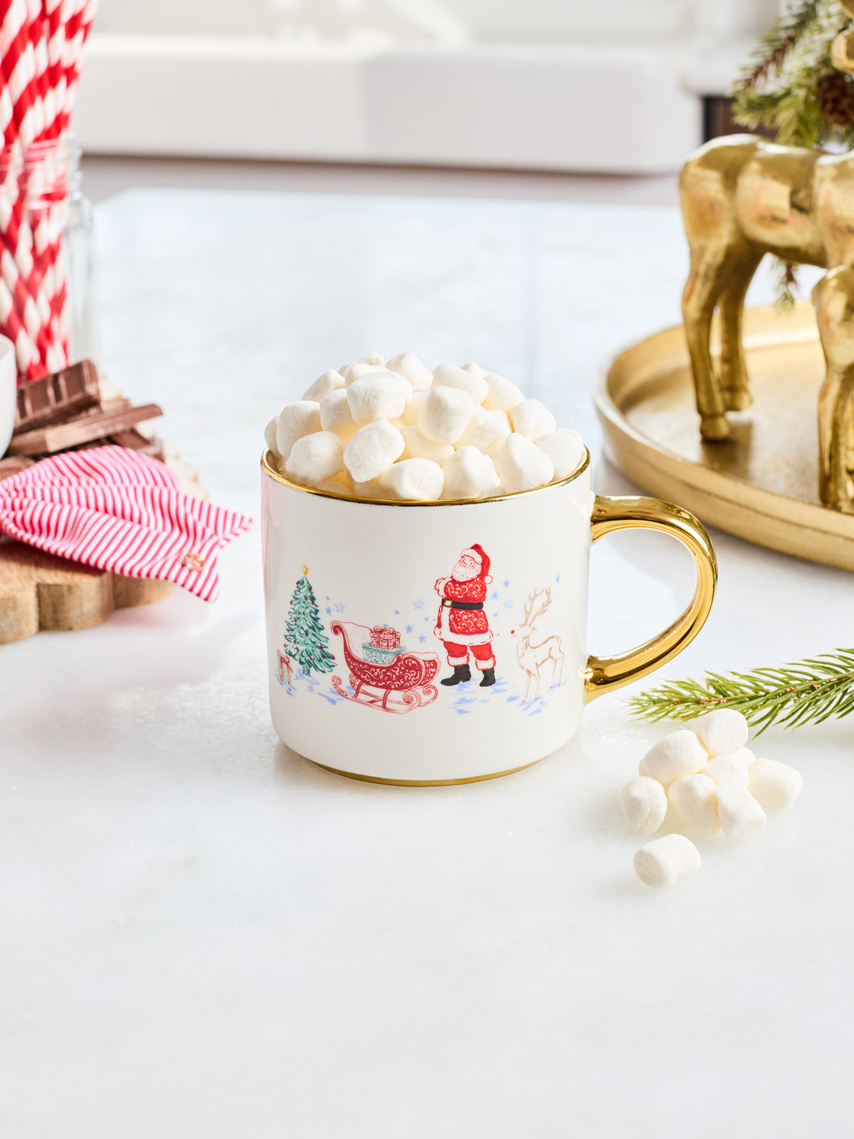 A white ceramic mug with a gold handle and rim, decorated with a festive Christmas scene featuring Santa Claus, a reindeer, and a tree. The mug is filled with white marshmallows and styled on a light tabletop with holiday props.
