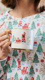 A person wearing a festive holiday-themed long-sleeve top holds a white ceramic mug with a gold rim and handle. The top features an all-over print of Santa Claus, sledding, reindeer, and Christmas trees in red, green, and brown on a white background.