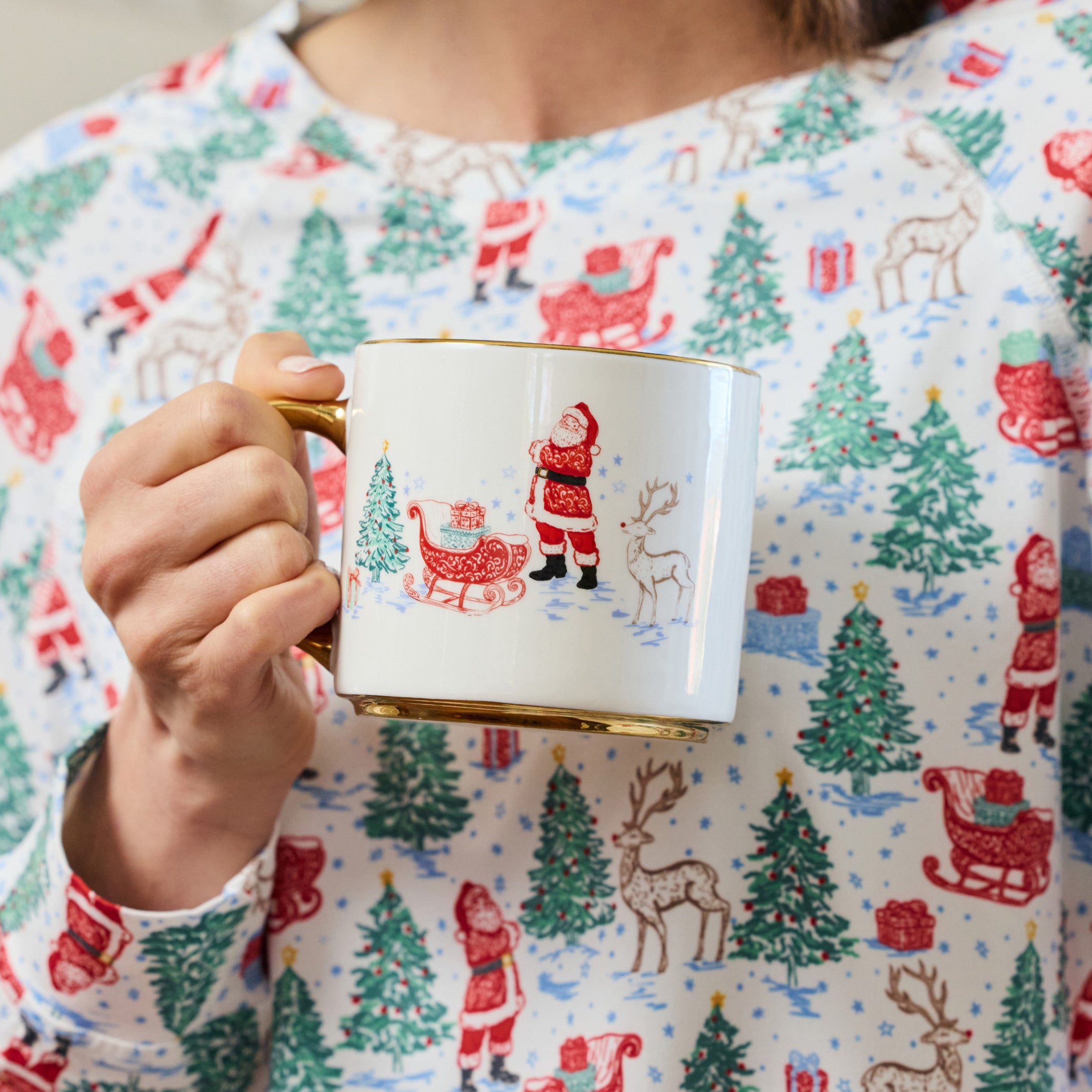 A person wearing a festive holiday-themed long-sleeve top holds a white ceramic mug with a gold rim and handle. The top features an all-over print of Santa Claus, sledding, reindeer, and Christmas trees in red, green, and brown on a white background.
