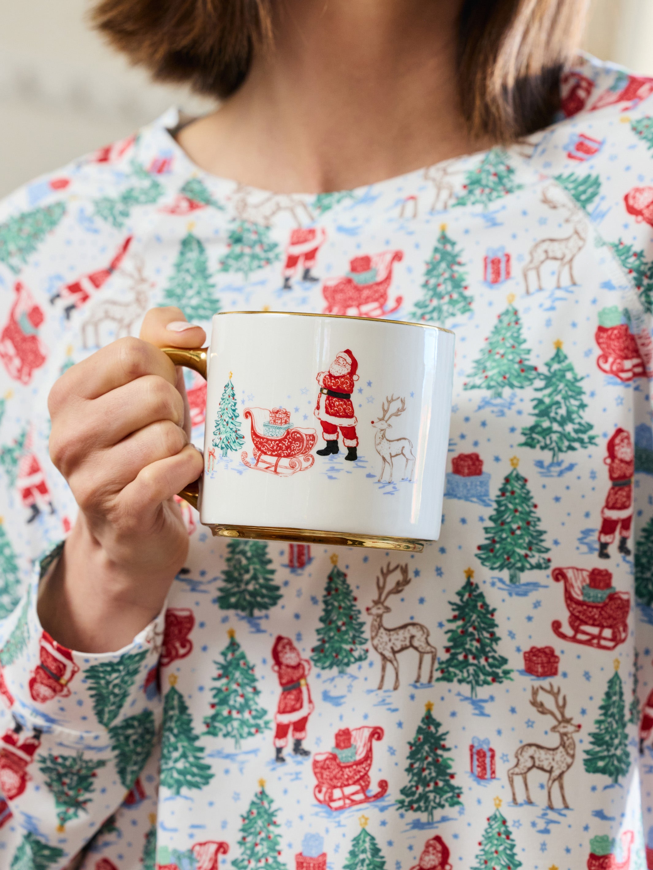 A person wearing a festive holiday-themed long-sleeve top holds a white ceramic mug with a gold rim and handle. The top features an all-over print of Santa Claus, sledding, reindeer, and Christmas trees in red, green, and brown on a white background.
