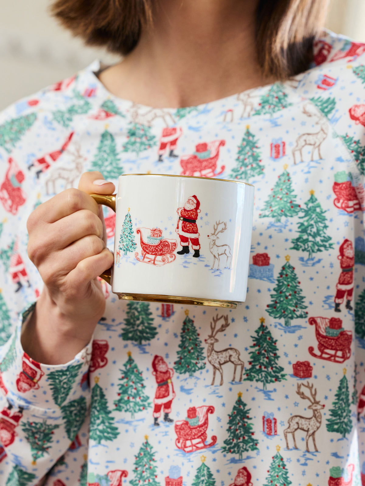 A person wearing a festive holiday-themed long-sleeve top holds a white ceramic mug with a gold rim and handle. The top features an all-over print of Santa Claus, sledding, reindeer, and Christmas trees in red, green, and brown on a white background.