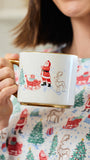A person holding a white ceramic mug with a gold handle and base, decorated with a Christmas scene featuring Santa Claus, a sleigh, a tree, and a reindeer. The person is wearing a white top with a red and green Christmas print.