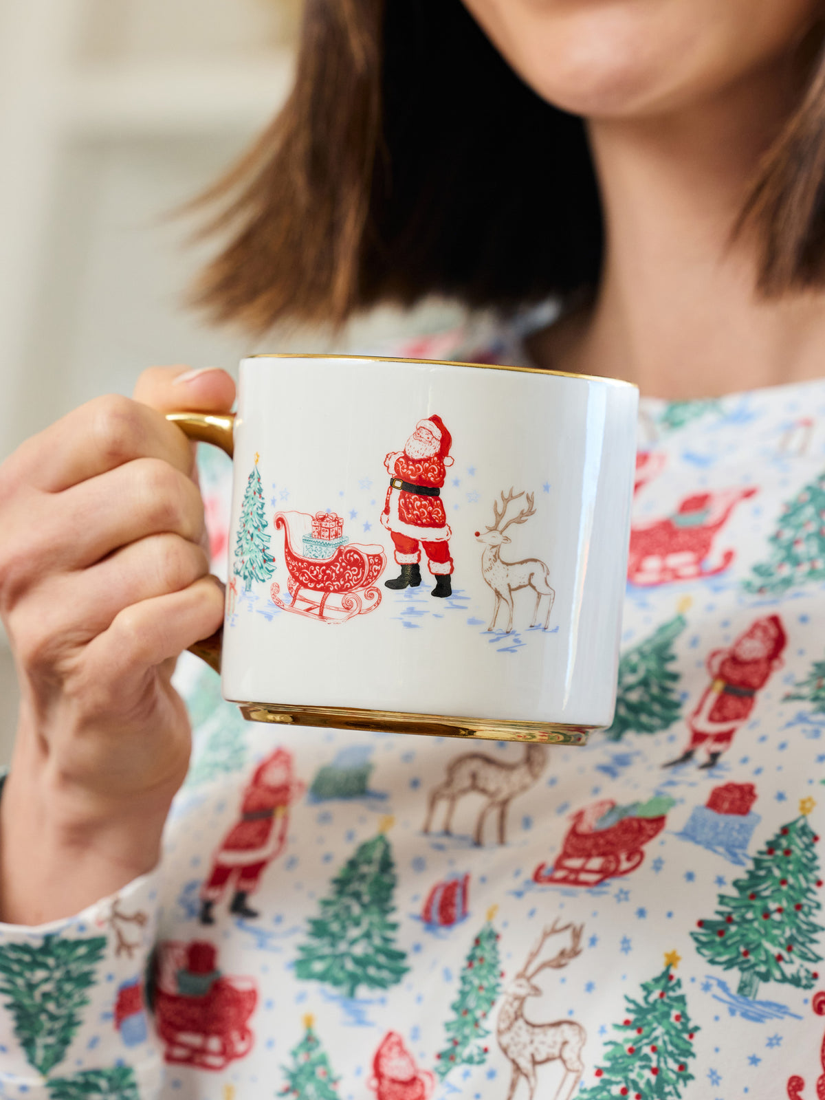A person holding a white ceramic mug with a gold handle and base, decorated with a Christmas scene featuring Santa Claus, a sleigh, a tree, and a reindeer. The person is wearing a white top with a red and green Christmas print.