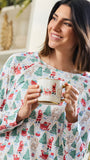 A woman wearing a festive holiday pajama top with a print of Santa Claus, reindeer, and Christmas trees, holding a white mug with gold trim.