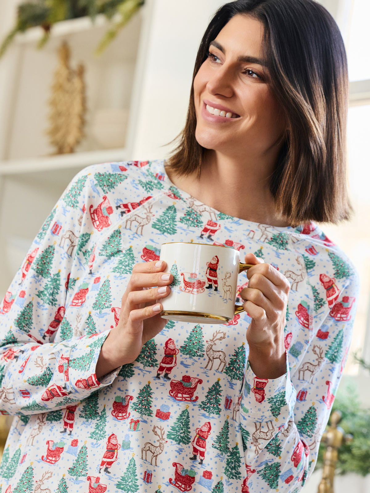 A woman wearing a festive holiday pajama top with a print of Santa Claus, reindeer, and Christmas trees, holding a white mug with gold trim.
