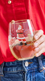 A person in a red shirt and blue jeans holds a clear stemless wine glass decorated with a Christmas Santa Claus and sleigh illustration. The glass is partially filled with a light-colored beverage.