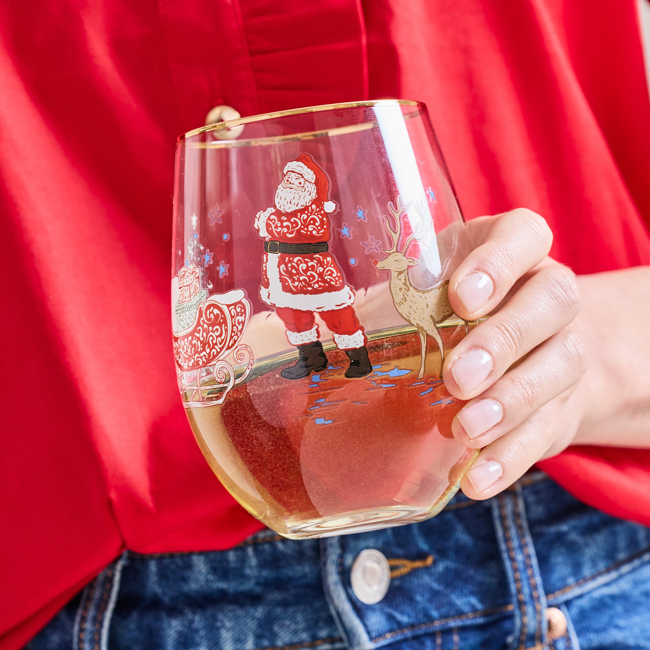 A person in a red shirt and blue jeans holds a clear stemless wine glass decorated with a Christmas Santa Claus and sleigh illustration. The glass is partially filled with a light-colored beverage.