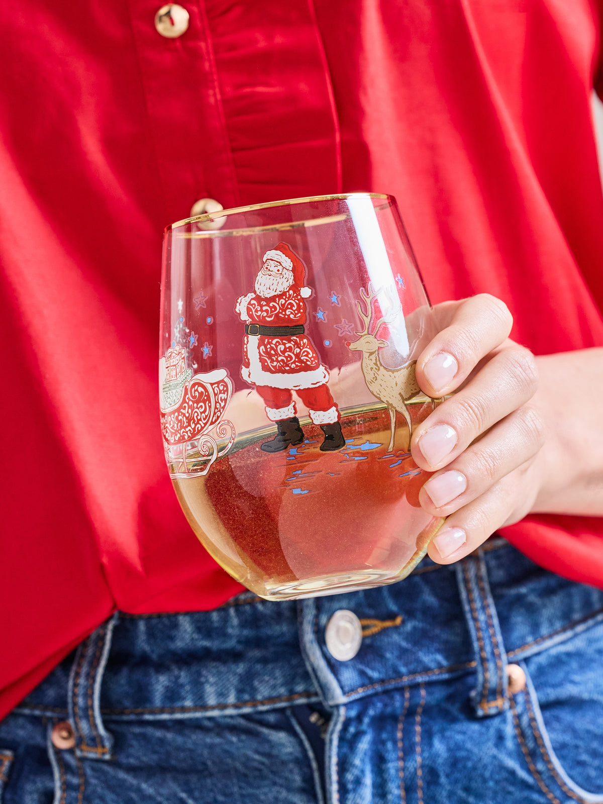 A person in a red shirt and blue jeans holds a clear stemless wine glass decorated with a Christmas Santa Claus and sleigh illustration. The glass is partially filled with a light-colored beverage.