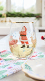 A clear stemless wine glass decorated with a festive Christmas illustration of Santa Claus, a sleigh, and a deer, shown on a dining table with a patterned napkin and plates.
