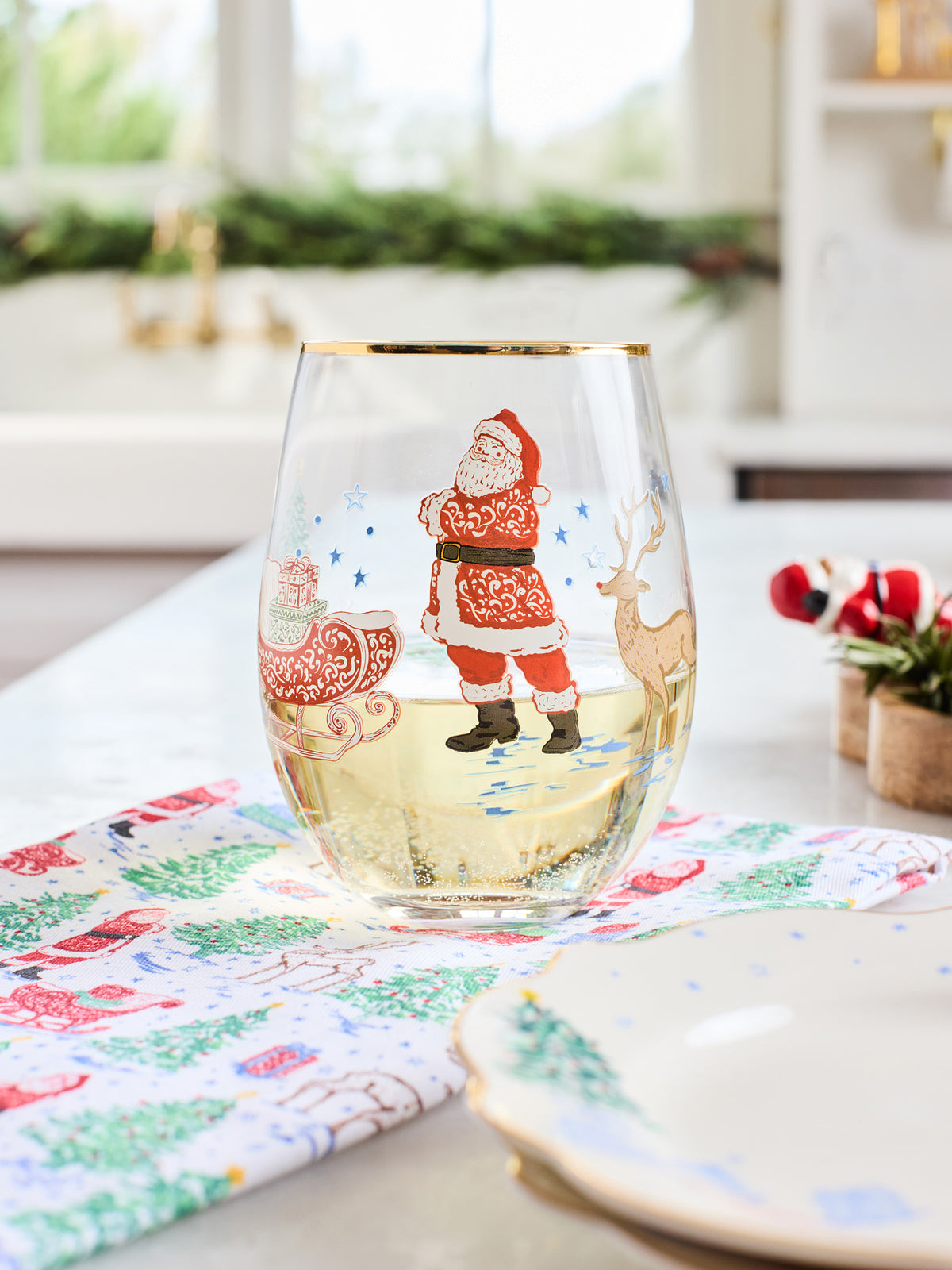 A clear stemless wine glass decorated with a festive Christmas illustration of Santa Claus, a sleigh, and a deer, shown on a dining table with a patterned napkin and plates.