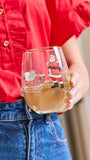 A person in a red shirt and blue jeans holds a clear stemless wine glass decorated with a Christmas Santa Claus and sleigh illustration. The glass is partially filled with a light-colored beverage.
