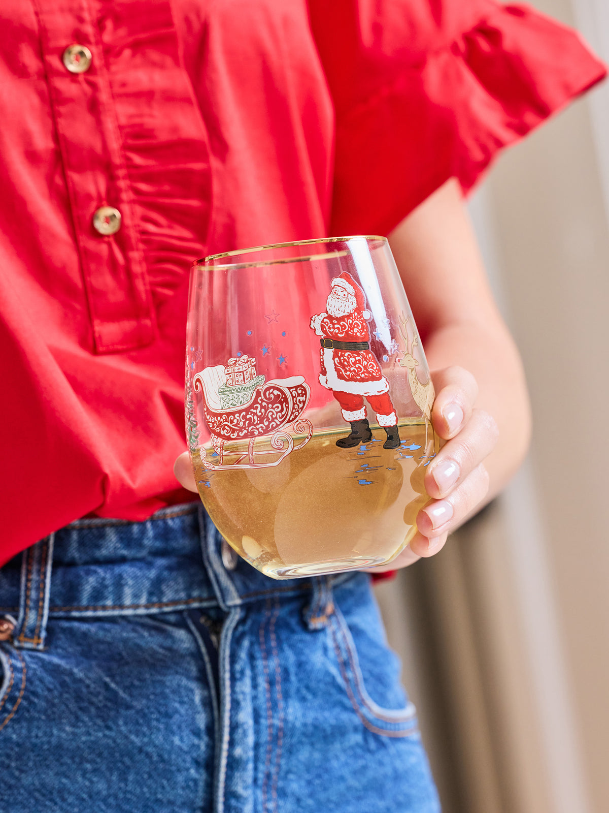 A person in a red shirt and blue jeans holds a clear stemless wine glass decorated with a Christmas Santa Claus and sleigh illustration. The glass is partially filled with a light-colored beverage.