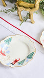 A scalloped ceramic holiday plate with a Santa Claus and reindeer design, styled on a white tablecloth with a red runner, greenery, and a gold reindeer figurine.