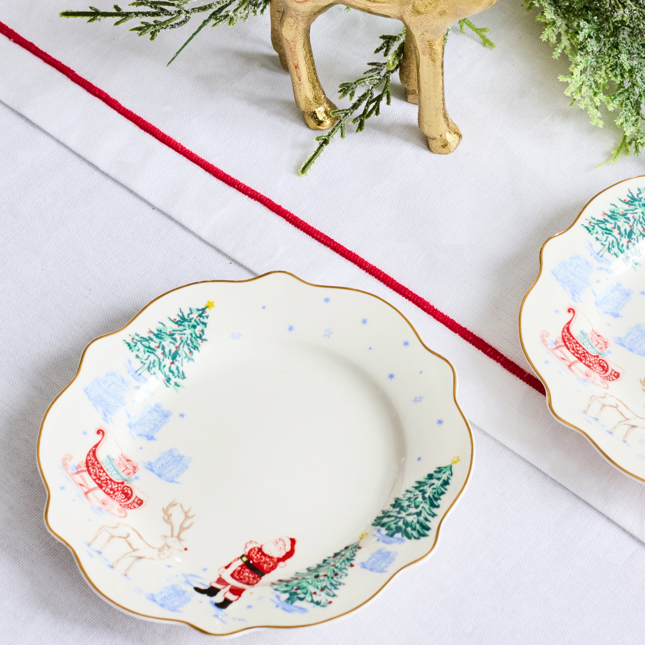A scalloped ceramic holiday plate with a Santa Claus and reindeer design, styled on a white tablecloth with a red runner, greenery, and a gold reindeer figurine.