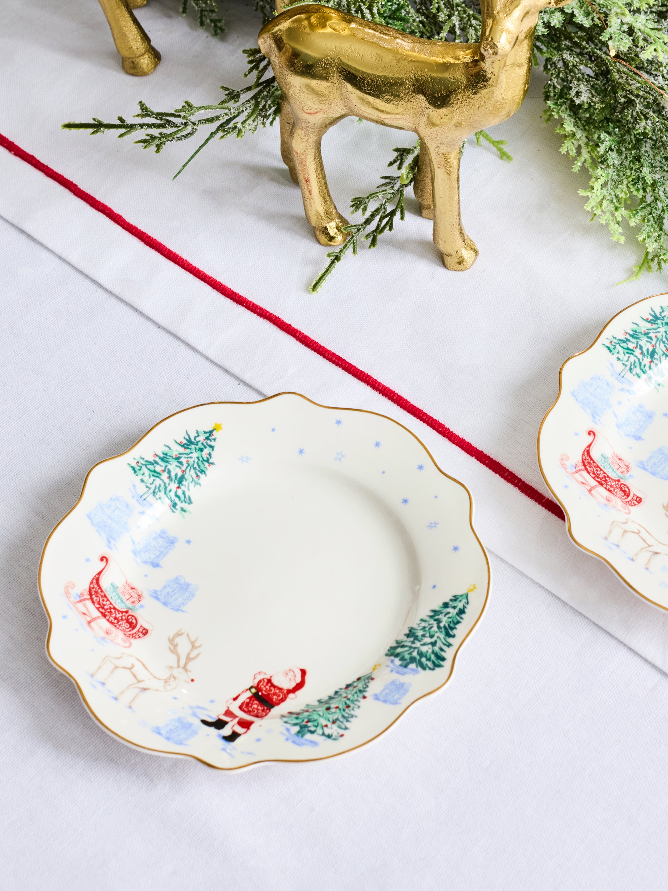 A scalloped ceramic holiday plate with a Santa Claus and reindeer design, styled on a white tablecloth with a red runner, greenery, and a gold reindeer figurine.