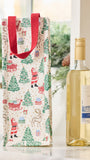 A Christmas-themed gift bag with a red handle stands next to a bottle of white wine on a light countertop. The bag has a cream background with a festive print of Santa Claus, reindeer, Christmas trees, sleighs, and gifts.